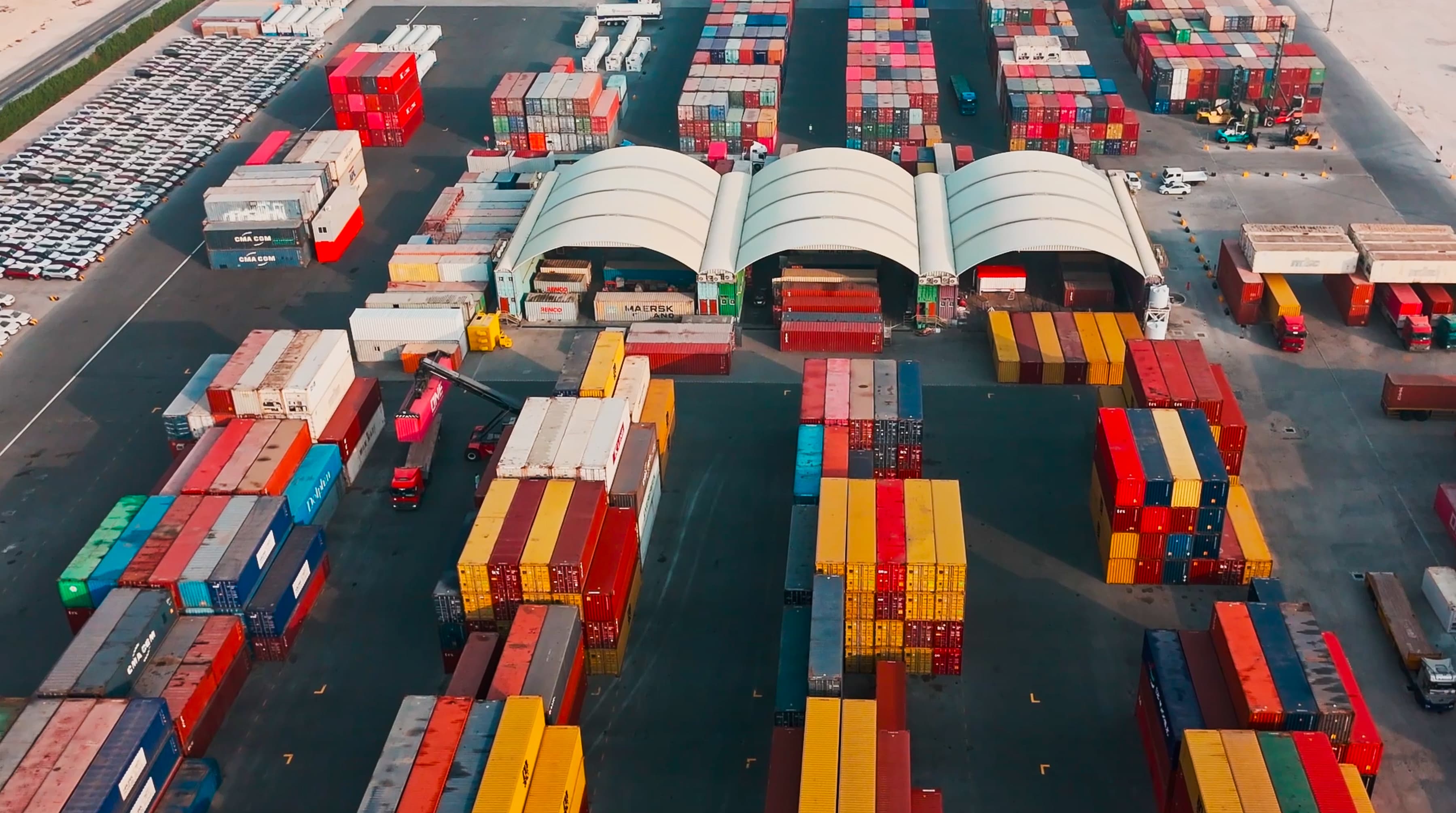 Aerial view of a busy shipping yard filled with colorful stacked containers and trucks, with parked cars in the background.