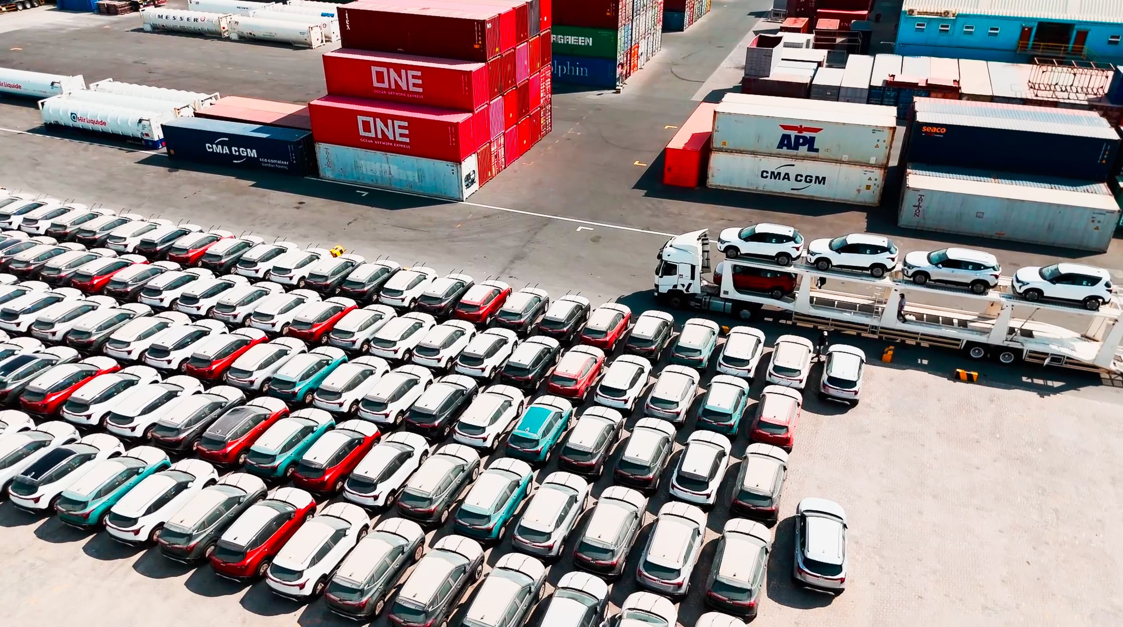 Aerial view of a parking lot filled with various colored cars, with a truck unloading vehicles near shipping containers.