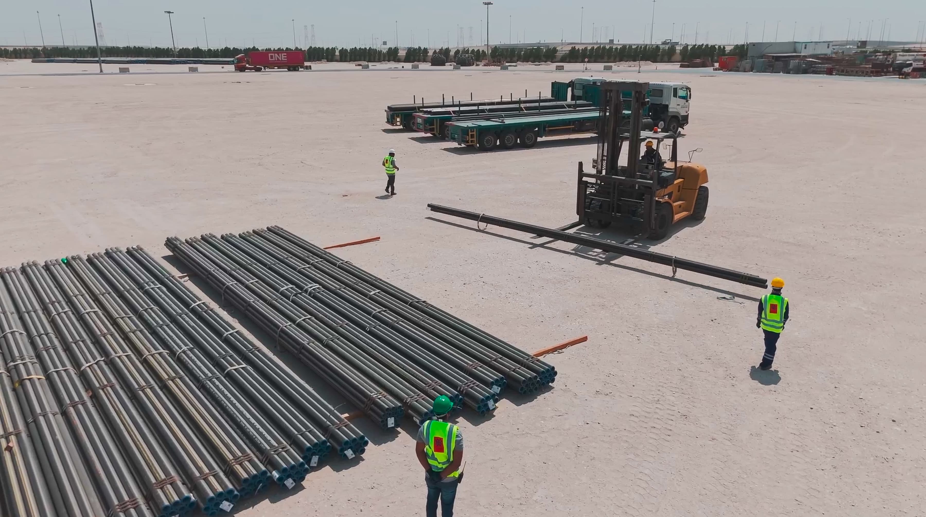 Two workers in safety vests oversee a forklift unloading steel pipes on a vast, empty construction site. Trucks are in the background.