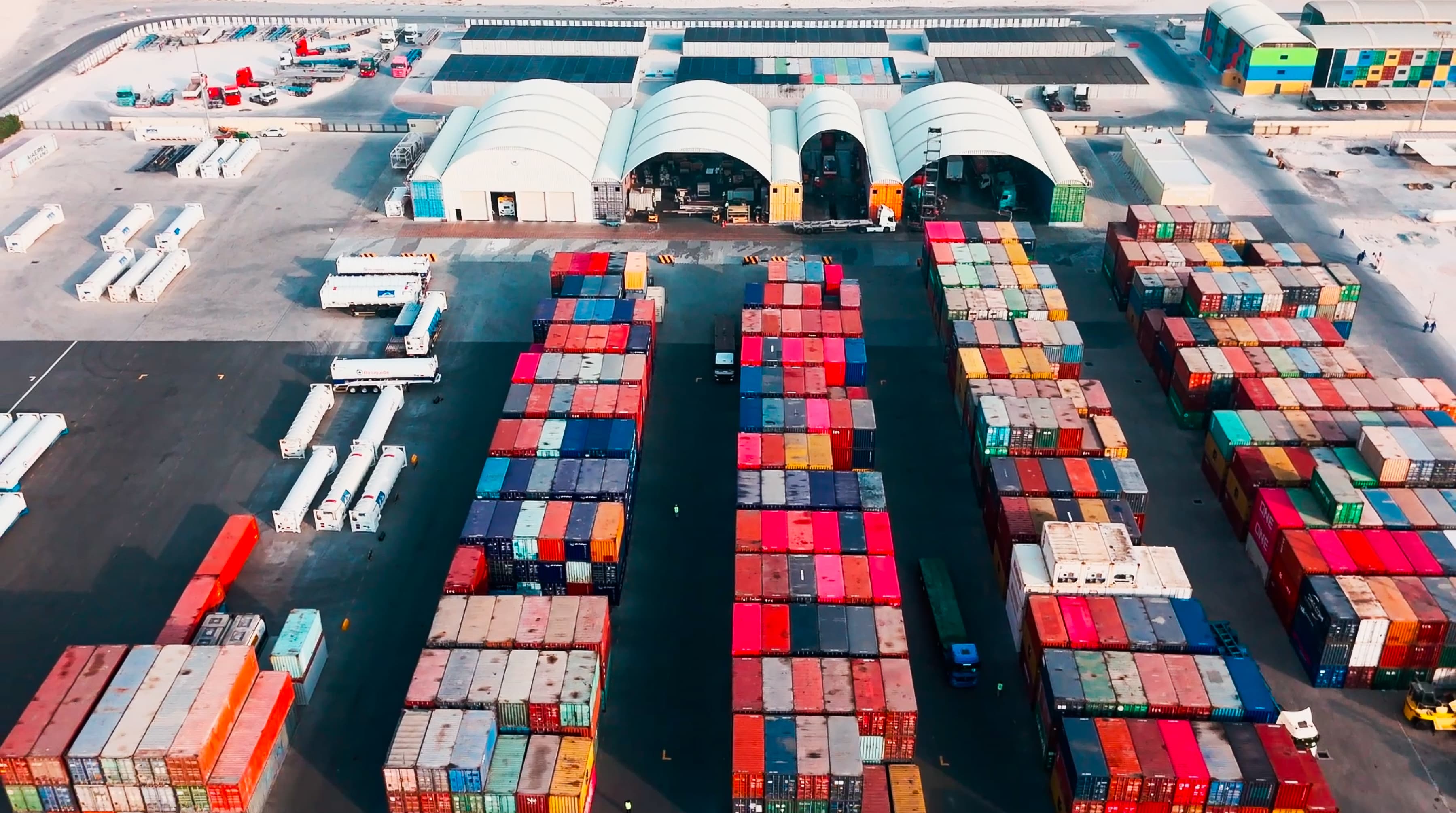 Aerial view of a busy shipping yard filled with colorful cargo containers and trucks, with storage facilities in the background.