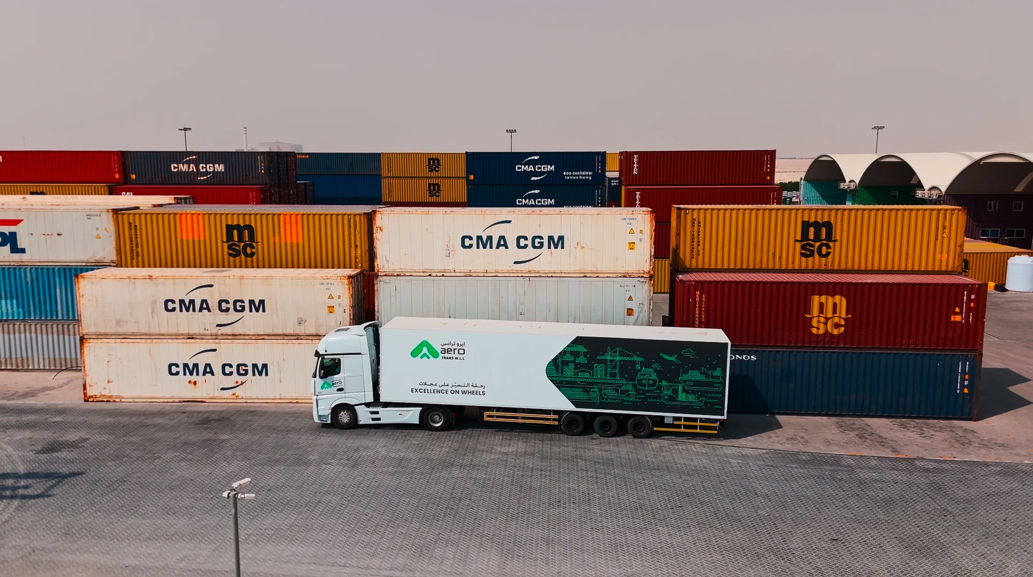 A truck with a branded trailer parked beside stacked shipping containers in a logistics yard under a hazy sky.