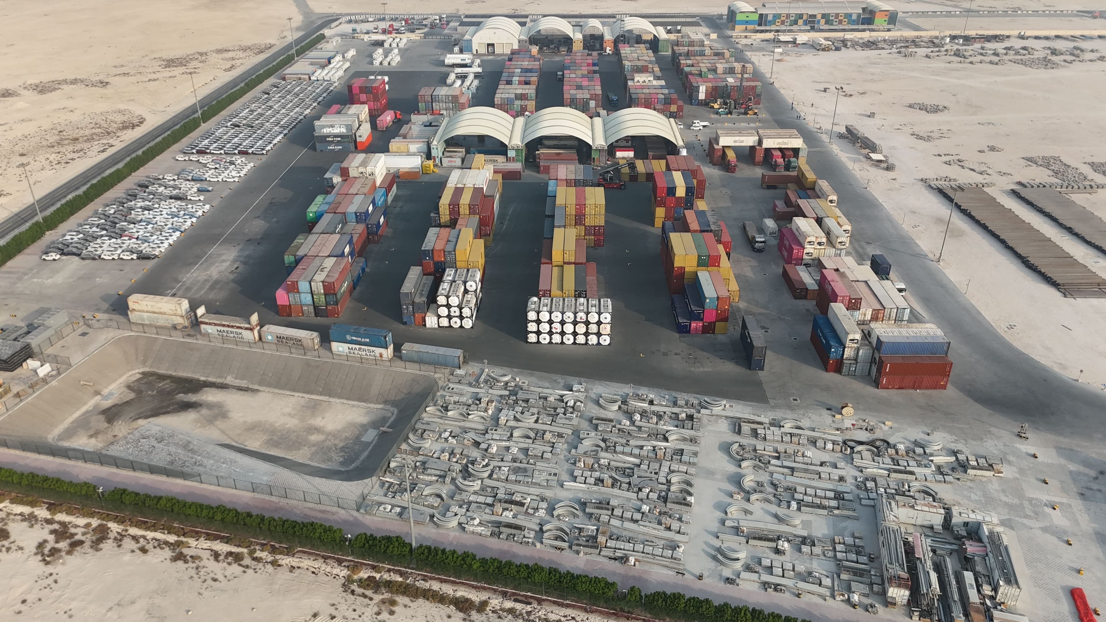 Ariel view of a shipping container yard with stacked containers, large white tents, and industrial materials organized on a paved lot in a desert area.