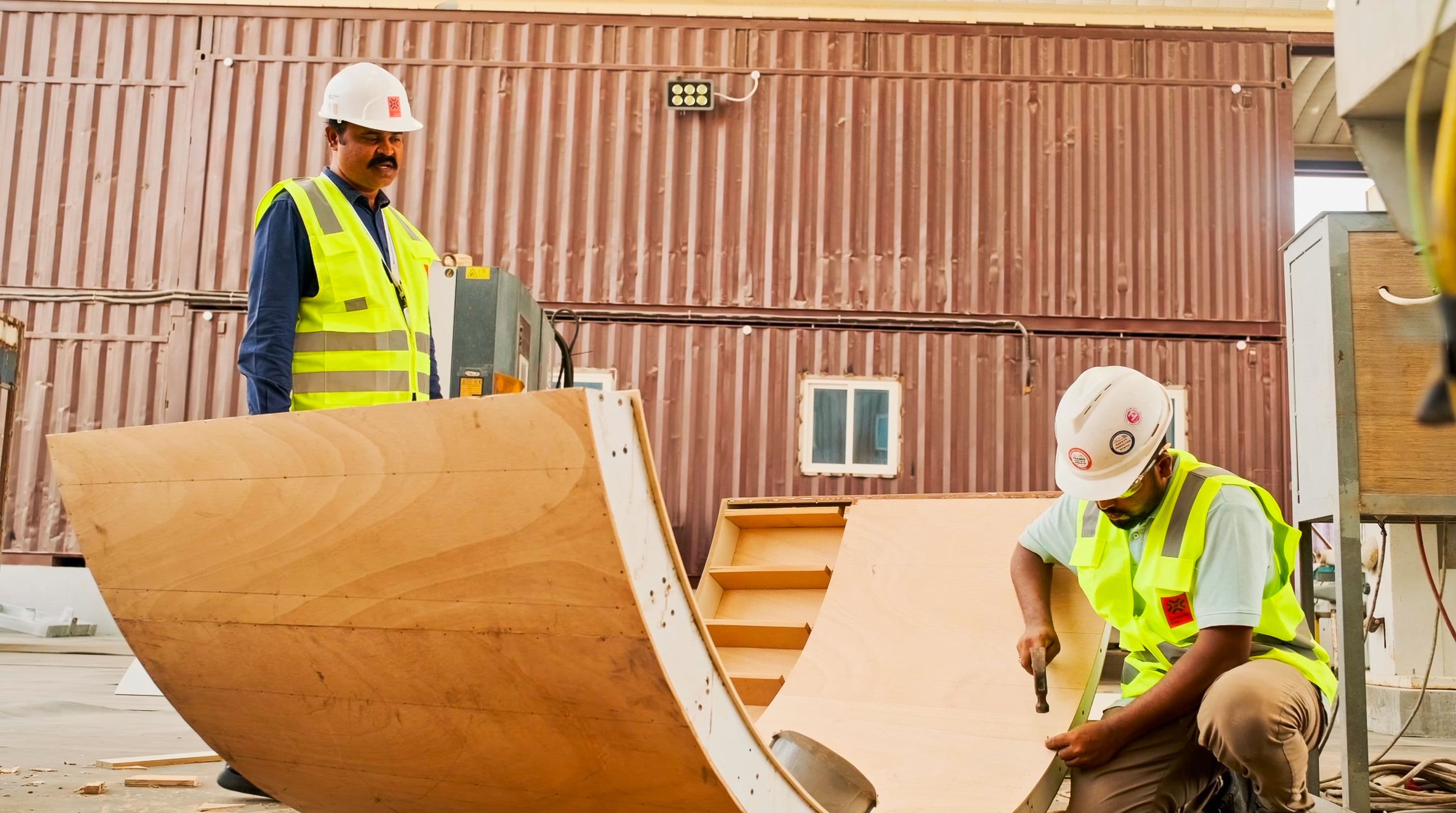 Two construction workers in safety gear and vests work on assembling large wooden structures outdoors, with a metal building in the background.