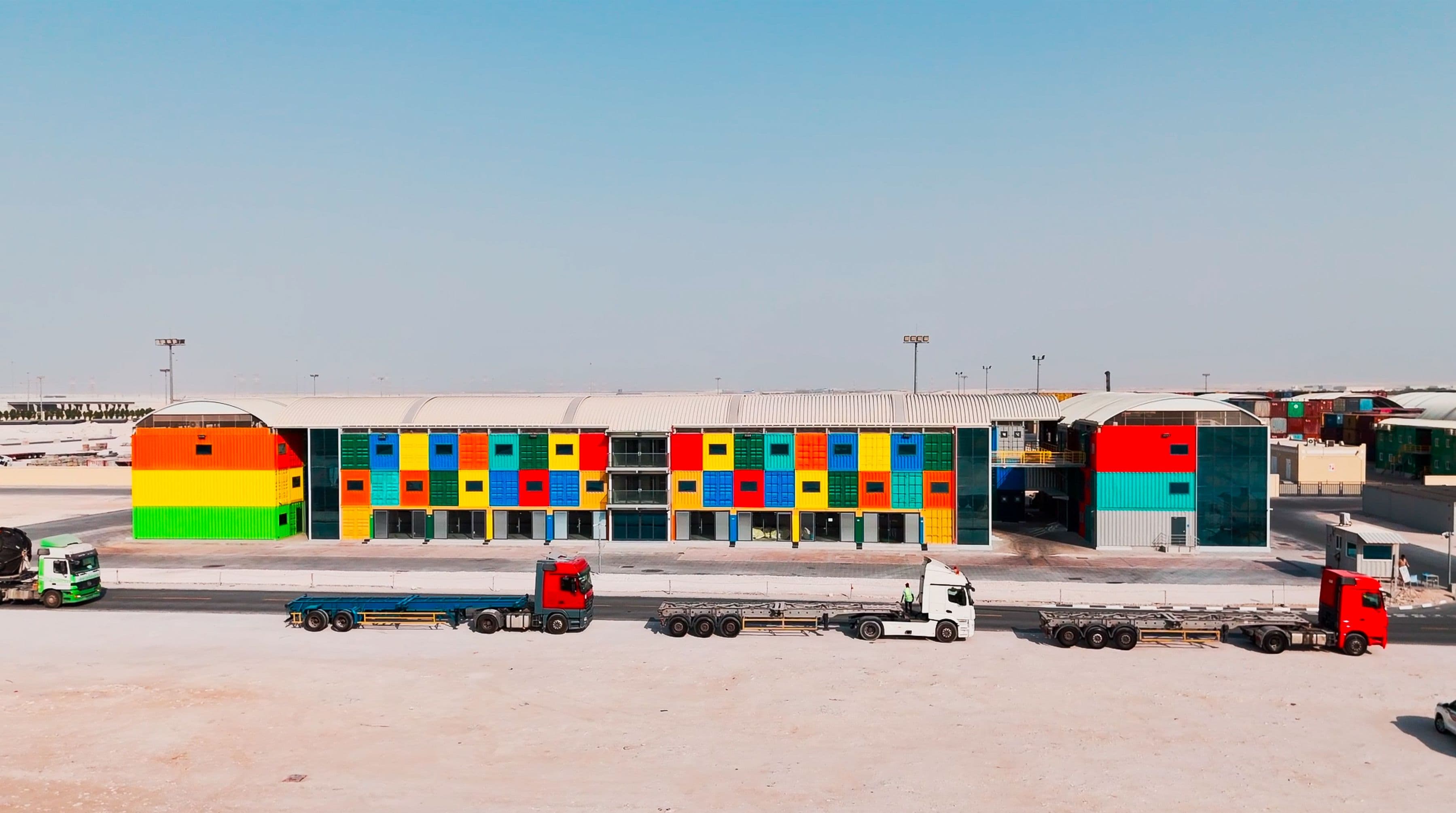 A vibrant, modern building with a colorful facade of red, blue, green, and yellow panels. Four large trucks are parked in front under a clear sky.