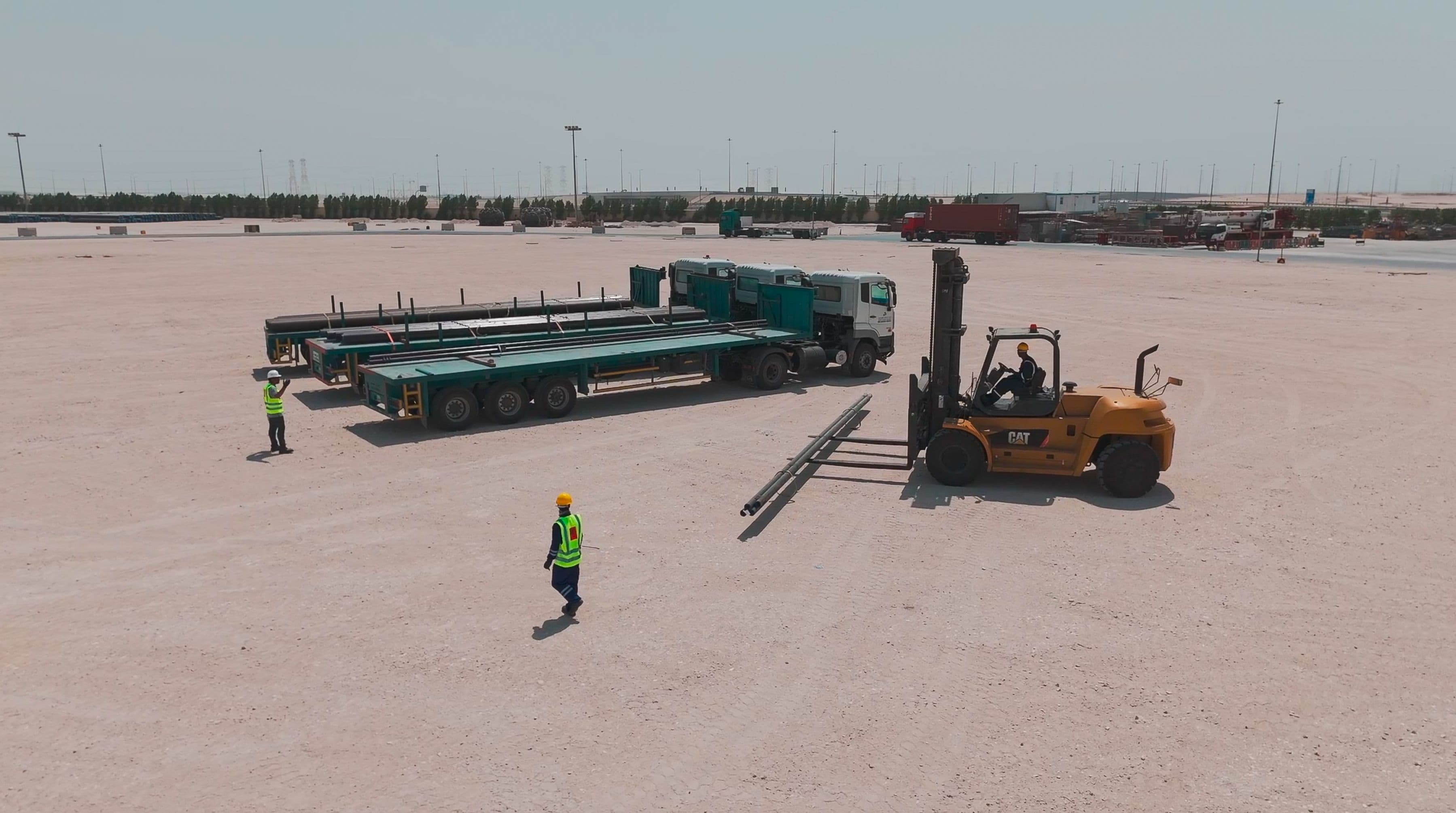 A construction site with two flatbed trucks and a forklift handling materials. Three workers in safety vests and helmets are present on the sandy terrain.