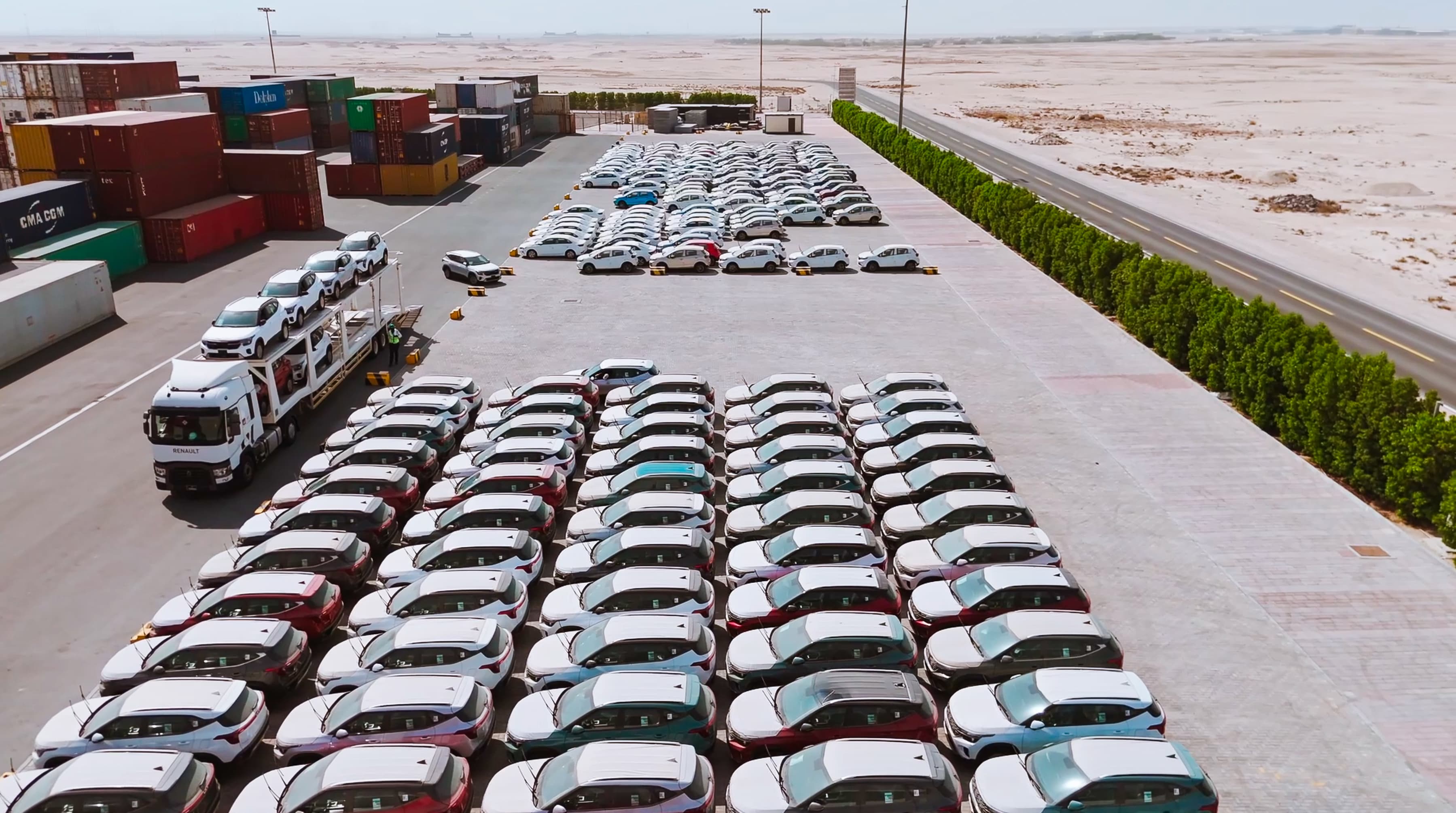 Overhead view of rows of white cars parked in a lot beside a desert road. Shipping containers are stacked nearby under a clear blue sky.