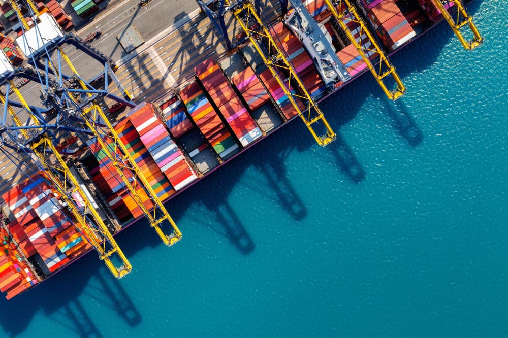Aerial view of a port with colorful shipping containers and cranes casting shadows on turquoise water.